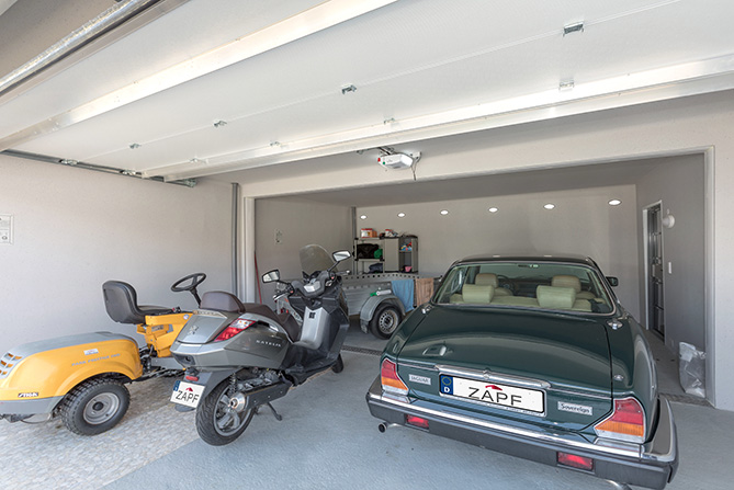 Eine großzügige Garage mit offenem Tor, in der sich ein grünes Oldtimer-Fahrzeug, ein gelber Aufsitzrasenmäher und ein grauer Motorroller befinden. Im Hintergrund sind ein Regal mit Werkzeugen und ein Anhänger zu sehen. Die Garage wirkt aufgeräumt und gut