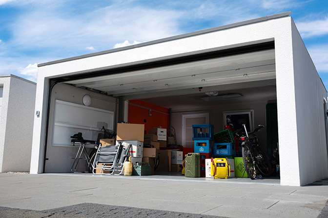 Eine Garage mit geöffnetem Tor, gefüllt mit verschiedenen Gegenständen wie Fahrrädern, Kartons, Klappstühlen und Werkzeug. Der Boden ist teilweise blockiert, und die Gegenstände sind unorganisiert verteilt.