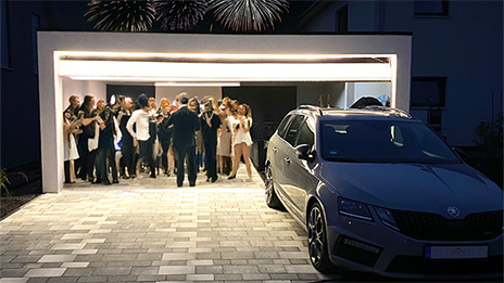 Moderne Garage mit beleuchtetem Tor, geparktes Auto im Vordergrund und festliche Partygesellschaft im Hintergrund, begleitet von Feuerwerk