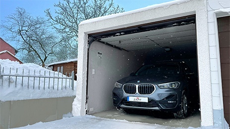 Schneebedeckte Garage mit geöffnetem Tor, in der ein BMW geparkt ist, umgeben von einer winterlichen Landschaft