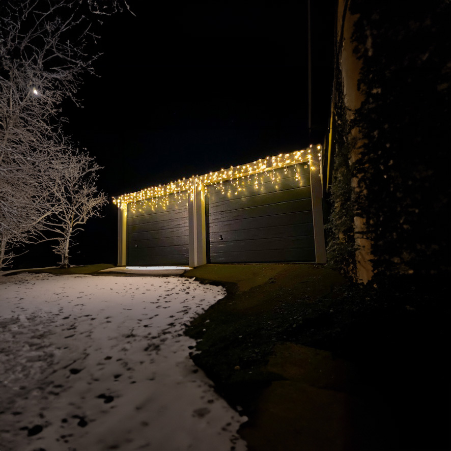 Garage mit schwarzer Tür, beleuchtet von warmweißen Lichterketten, im Schnee bedeckten Winterabend